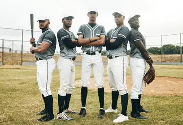 Five baseball players in gray uniforms pose confidently on a field, with one holding a bat, ready for the game — a perfect scene for an mlb players travel ball breakdown.